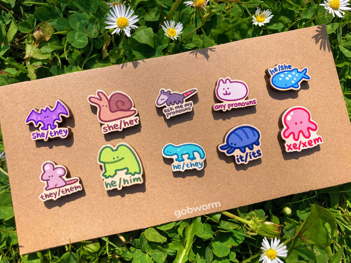 Set of colorful pronoun pins on a cardboard backing with grass and flowers in the background