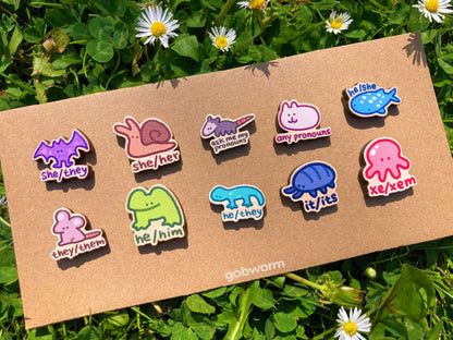 Set of colorful pronoun pins on a cardboard backing with grass and flowers in the background