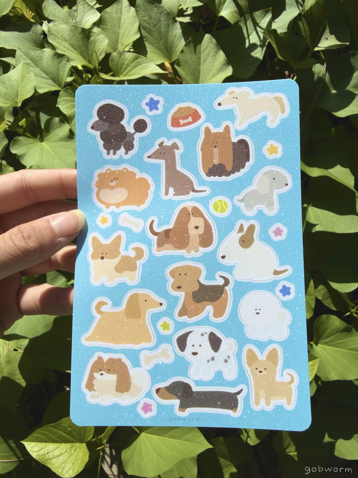 hand holding a sticker sheet featuring lineless illustrations of various dog breeds with a light blue background and glittery finish