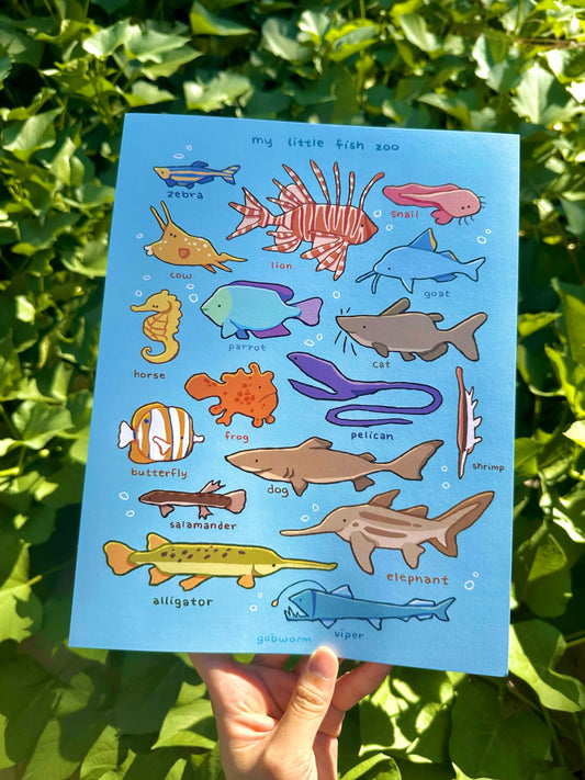 A hand holds a blue educational children's poster titled 'my little fish zoo' featuring colorful cartoon illustrations of different fish and marine life, set against a background of green outdoor foliage.