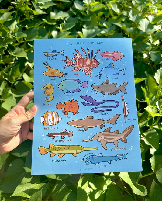 A hand holds a blue children's educational poster titled 'my little fish zoo' in front of green foliage. The poster features colorful illustrations of various fish and marine life with labels.