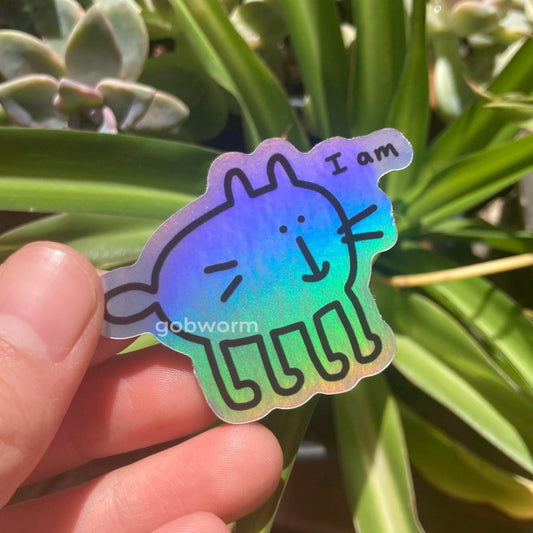 Holographic die-cut sticker with a cat design and text "i am" held in front of green plants